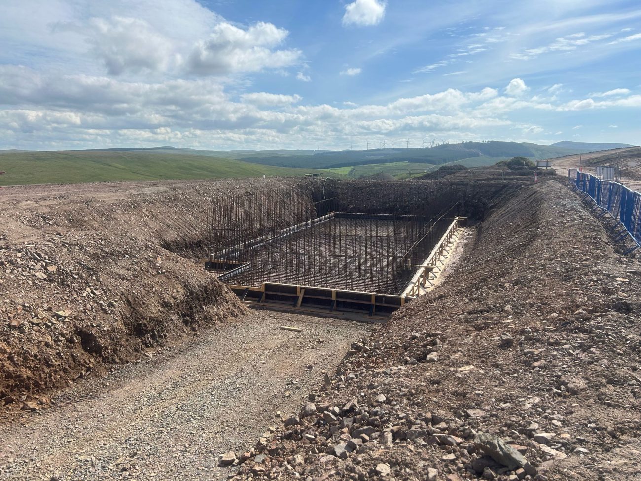 Reinforced concrete foundation slab being prepared within an excavated platform at Hagshaw Hill Windfarm, with wind turbines visible across the hillside in the background.